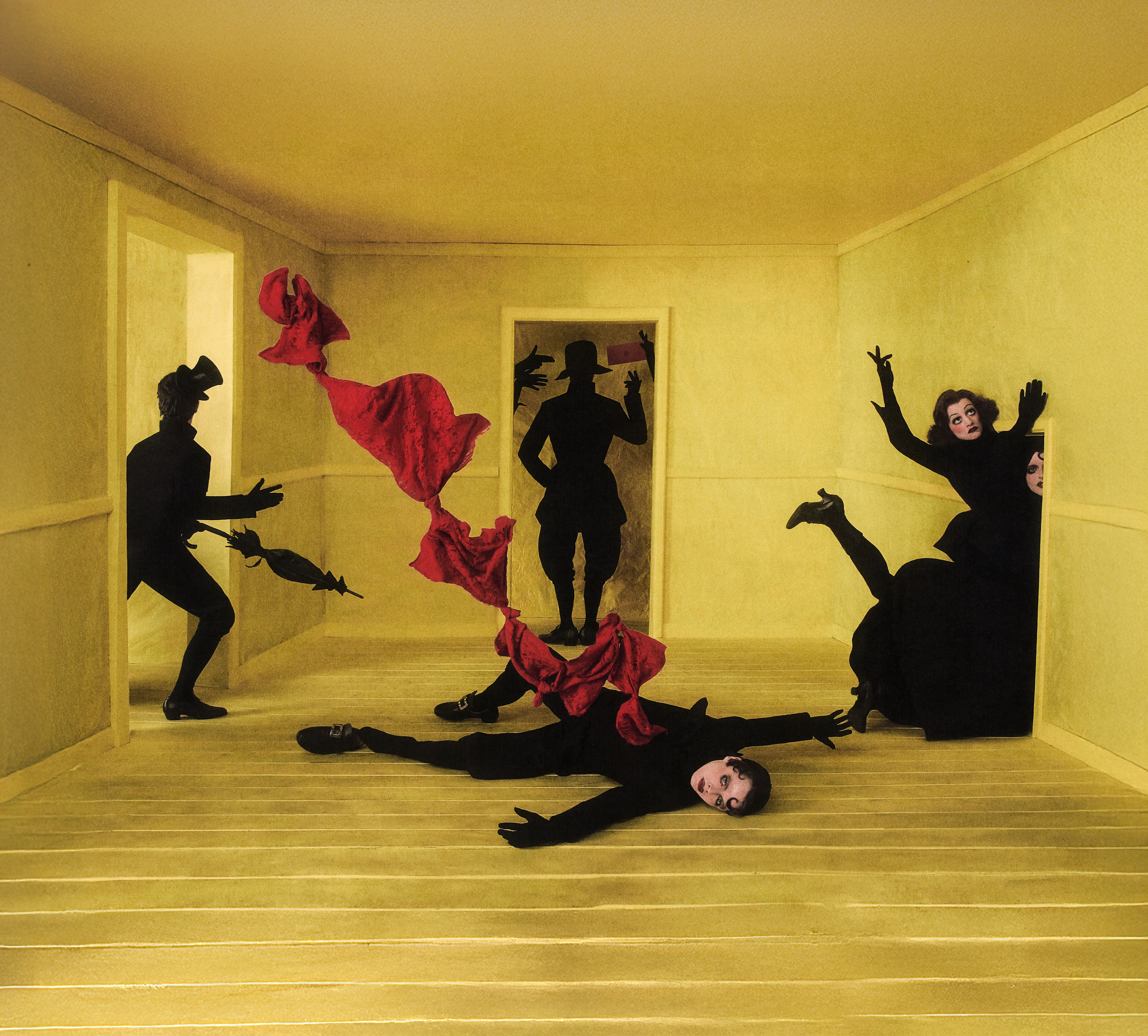 Surreal theatrical scene in an empty yellow room, representing the set design of Philipp Stölzl’s Un ballo in maschera. Six black-clad figures with dramatic 1920’s-inspired make-up appear frozen in dramatic poses around the room. One figure lies sprawled on the wooden yellow floor with a red piece of fabric emerging from his vest and twisting through the air at the centre. Other figures emerge from the doorways around the central character, creating a dreamlike, unsettling atmosphere.