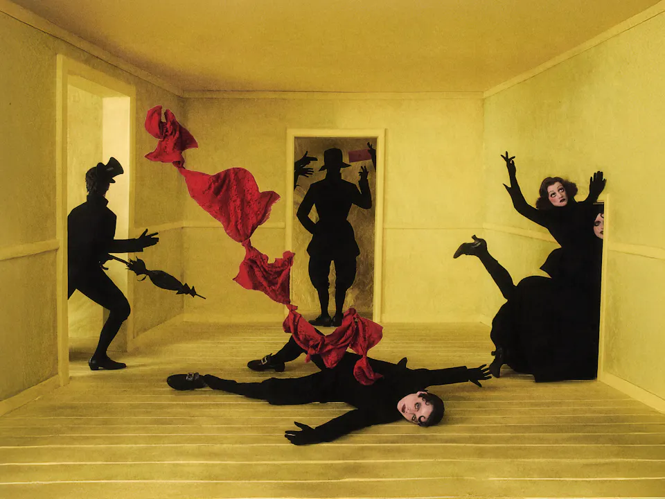 Surreal theatrical scene in an empty yellow room, representing the set design of Philipp Stölzl’s Un ballo in maschera. Six black-clad figures with dramatic 1920’s-inspired make-up appear frozen in dramatic poses around the room. One figure lies sprawled on the wooden yellow floor with a red piece of fabric emerging from his vest and twisting through the air at the centre. Other figures emerge from the doorways around the central character, creating a dreamlike, unsettling atmosphere.