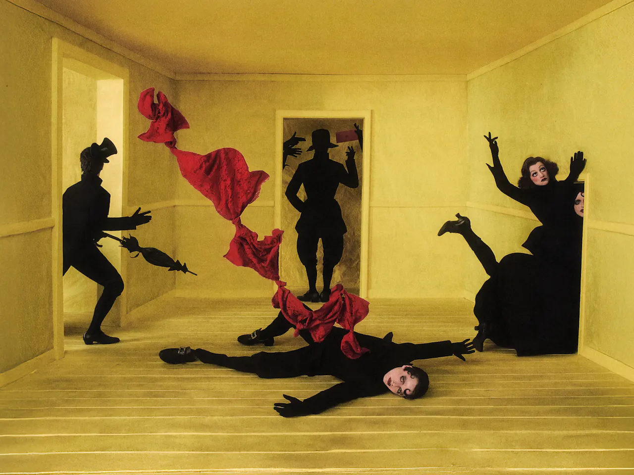 Surreal theatrical scene in an empty yellow room, representing the set design of Philipp Stölzl’s Un ballo in maschera. Six black-clad figures with dramatic 1920’s-inspired make-up appear frozen in dramatic poses around the room. One figure lies sprawled on the wooden yellow floor with a red piece of fabric emerging from his vest and twisting through the air at the centre. Other figures emerge from the doorways around the central character, creating a dreamlike, unsettling atmosphere.