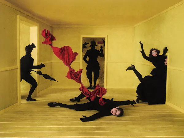 Surreal theatrical scene in an empty yellow room, representing the set design of Philipp Stölzl’s Un ballo in maschera. Six black-clad figures with dramatic 1920’s-inspired make-up appear frozen in dramatic poses around the room. One figure lies sprawled on the wooden yellow floor with a red piece of fabric emerging from his vest and twisting through the air at the centre. Other figures emerge from the doorways around the central character, creating a dreamlike, unsettling atmosphere.