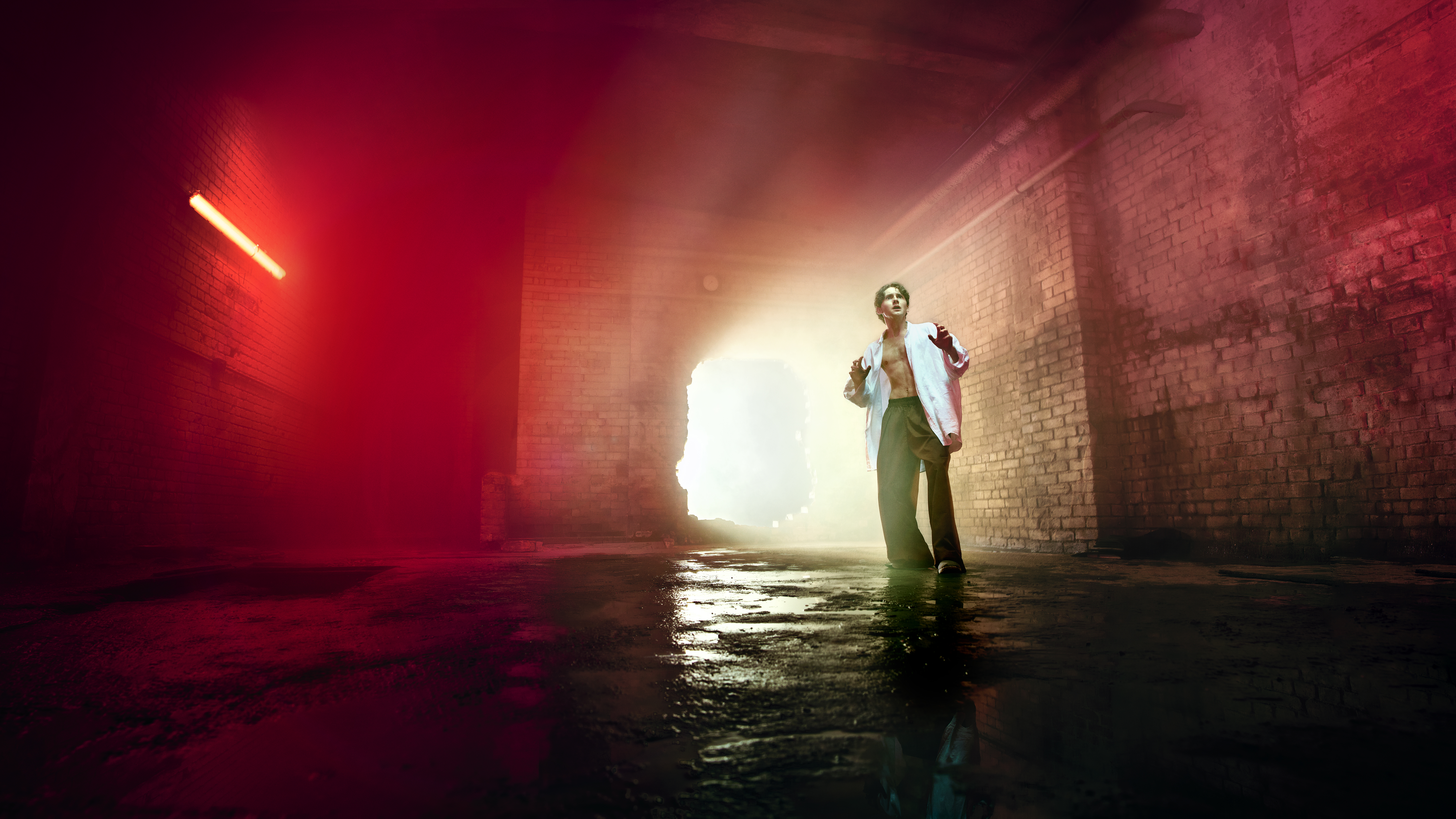 A young man representing Parsifal in Evgeny Titov’s Parsifal stands on a wet concrete floor in a claustrophobic, industrial brick interior. He wears an open white shirt and dark trousers. He looks upward with his hands raised slightly as bright light pours through a large opening behind him, illuminating the haze-filled space and fading into an eerie red glow at the edges of the frame.