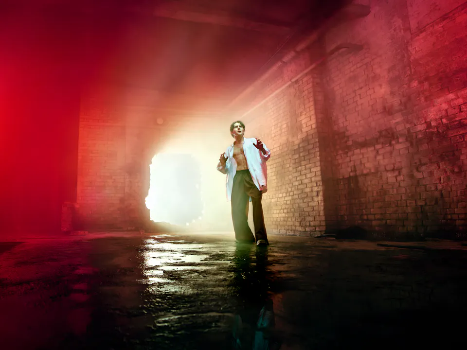 A young man representing Parsifal in Evgeny Titov’s Parsifal stands on a wet concrete floor in a claustrophobic, industrial brick interior. He wears an open white shirt and dark trousers. He looks upward with his hands raised slightly as bright light pours through a large opening behind him, illuminating the haze-filled space and fading into an eerie red glow at the edges of the frame.