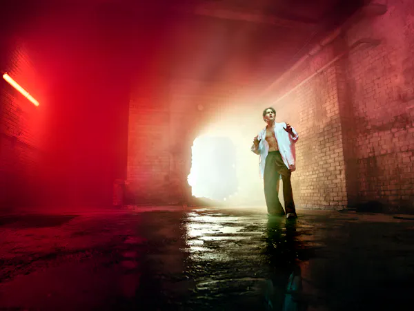 A young man representing Parsifal in Evgeny Titov’s Parsifal stands on a wet concrete floor in a claustrophobic, industrial brick interior. He wears an open white shirt and dark trousers. He looks upward with his hands raised slightly as bright light pours through a large opening behind him, illuminating the haze-filled space and fading into an eerie red glow at the edges of the frame.