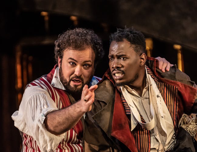 Two men in 19th-century outfits whisper together.