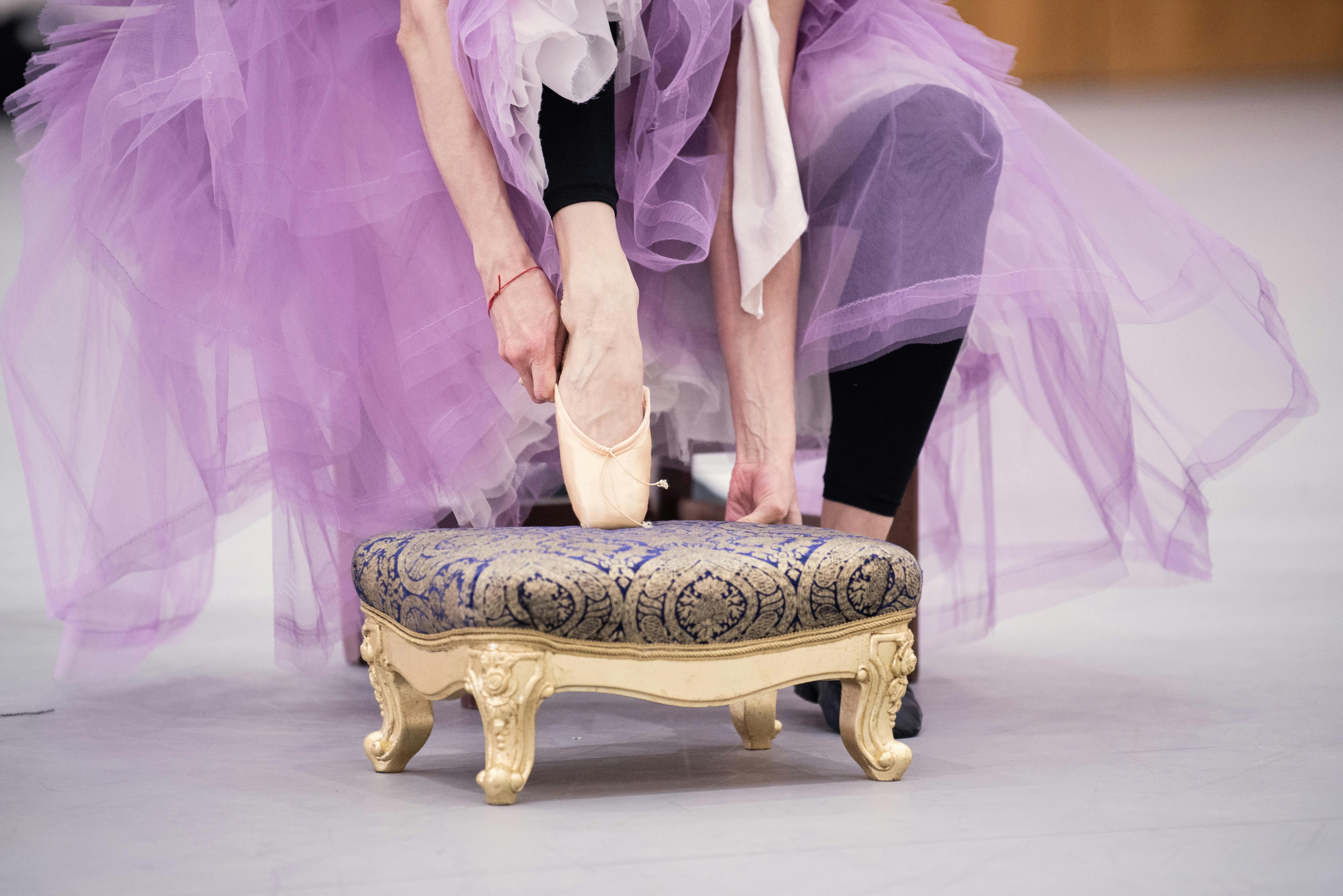 A dancer wearing a long purple tutu adjusts her pointe shoe, resting her foot on a small stool.