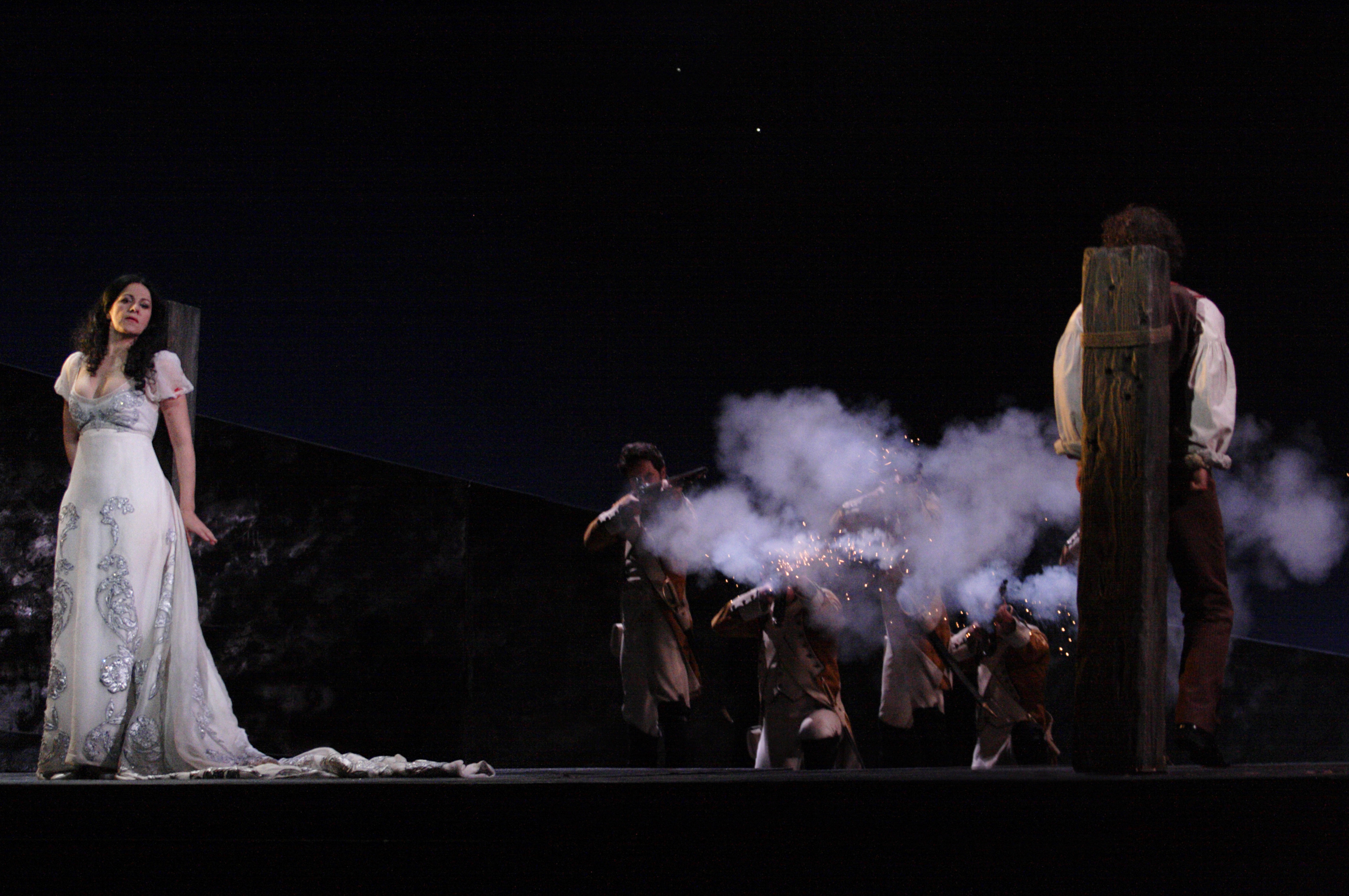 A woman in a white dress and faces forward and a man faces backwards. They are tied to stakes, waiting to be executed.
