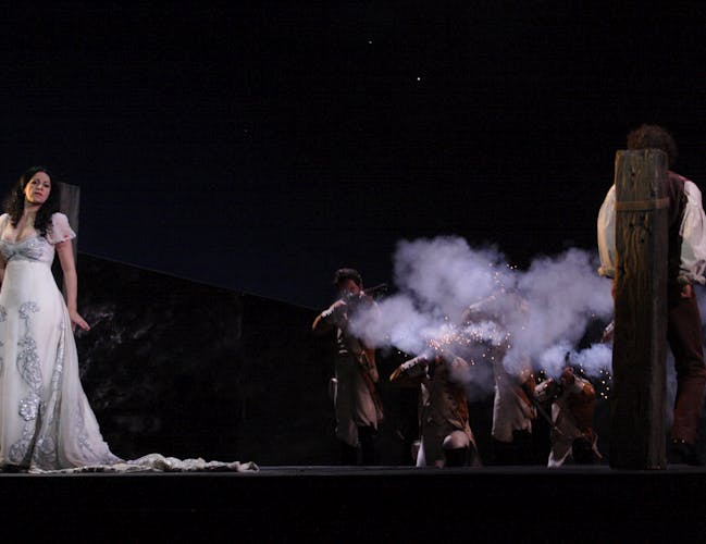 A woman in a white dress and faces forward and a man faces backwards. They are tied to stakes, waiting to be executed.