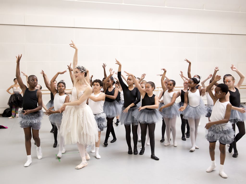 A group of school children in grey tutus copy an adult dancer dressed in a long white tutu dress in a white rehearsal room.