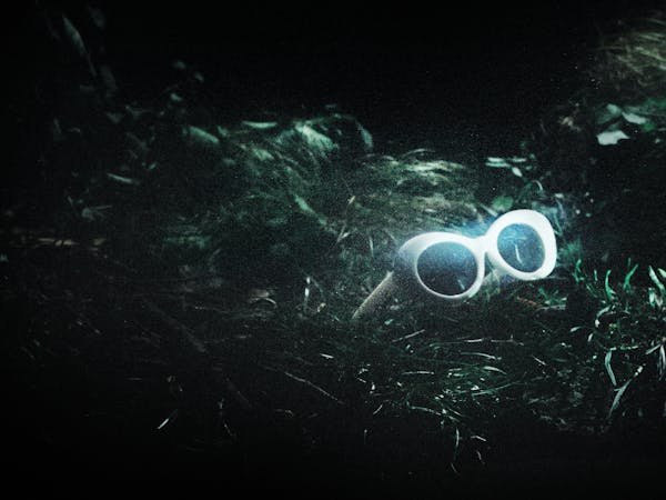 A pair of white 60s style framed sunglasses sit in amongst green grass. They represent the glasses worn by Kurt Cobain in The Royal Opera’s production of Last Days.