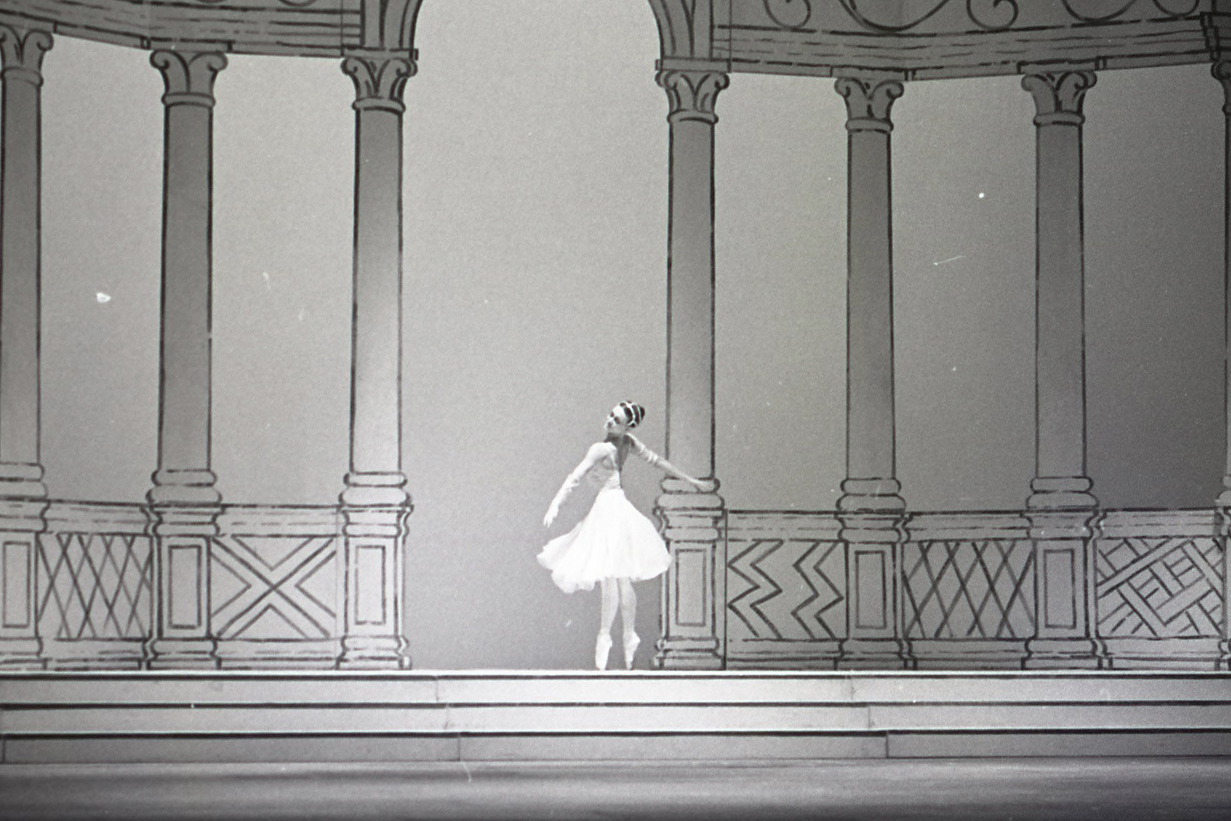 A dancer in a white dress stands on stage between a series of columns. (6 of 8)
