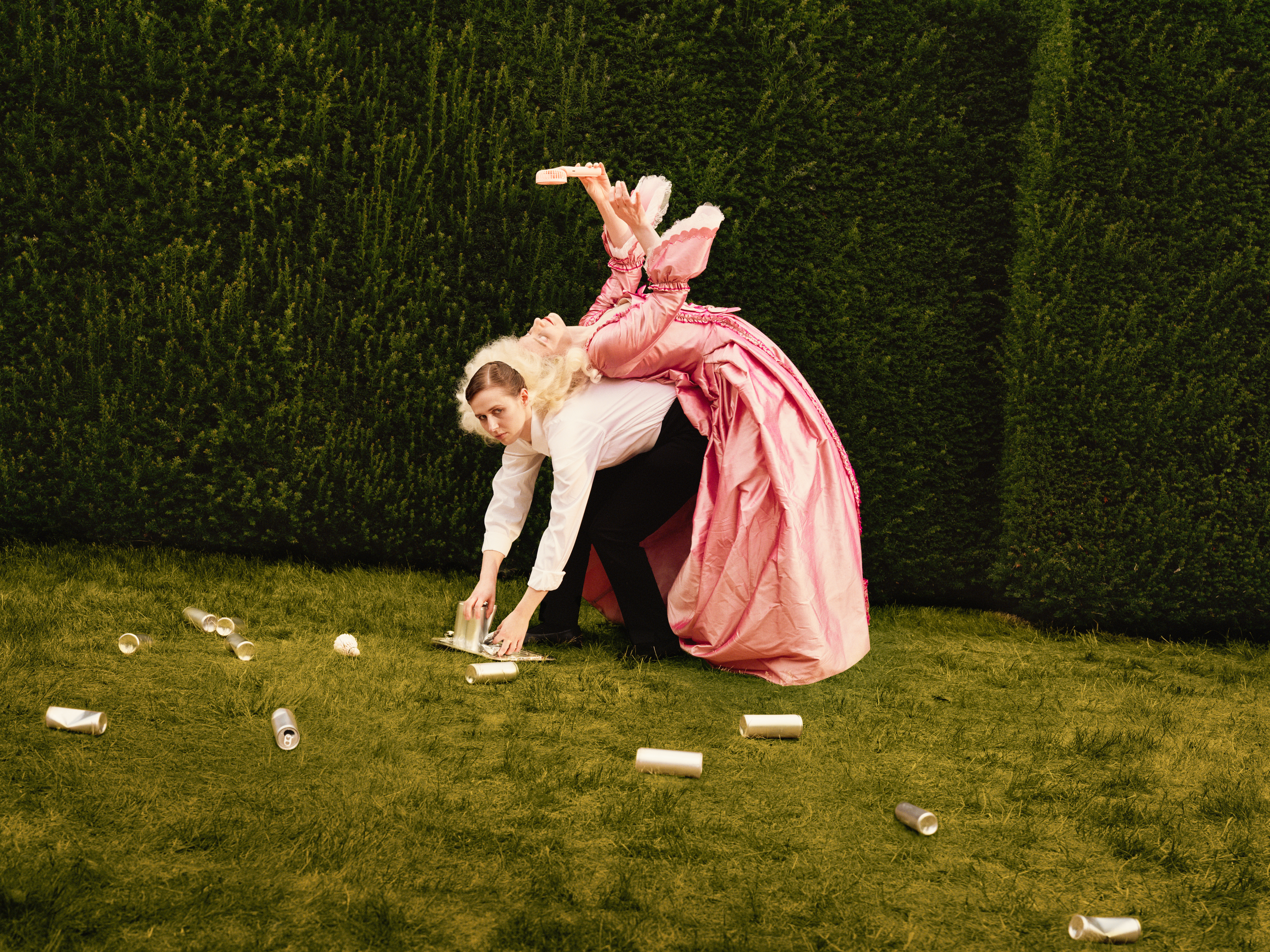 A woman in a maid costume with a white shirt and black trousers bends over on a lawn to collect empty cans from the ground, while a performer in an elaborate pink period costume lies across her back, holding an electric fan above her head.