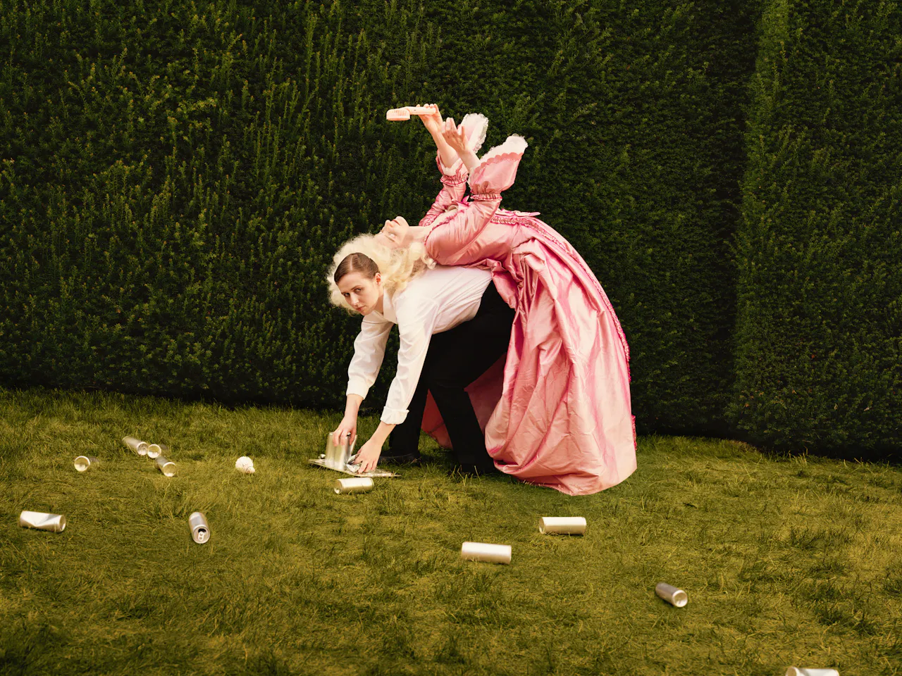 A woman in a maid costume with a white shirt and black trousers bends over on a lawn to collect empty cans from the ground, while a performer in an elaborate pink period costume lies across her back, holding an electric fan above her head.