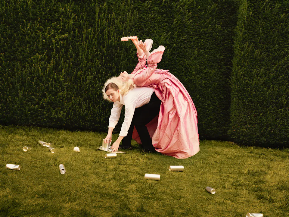 A woman in a maid costume with a white shirt and black trousers bends over on a lawn to collect empty cans from the ground, while a performer in an elaborate pink period costume lies across her back, holding an electric fan above her head.
