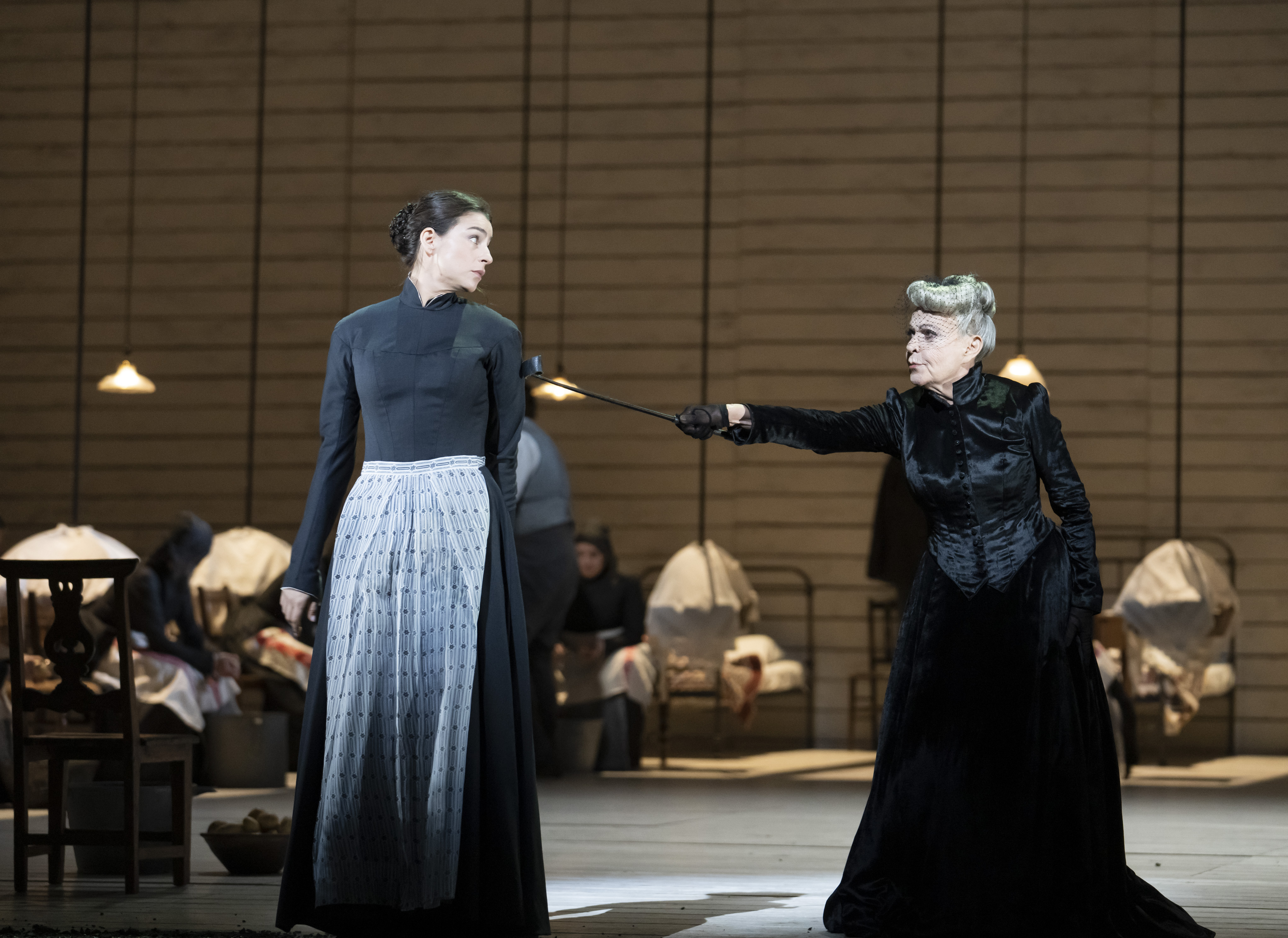 An old woman in a black dress points accusingly at a younger woman in a dark navy dress and white apron. 
