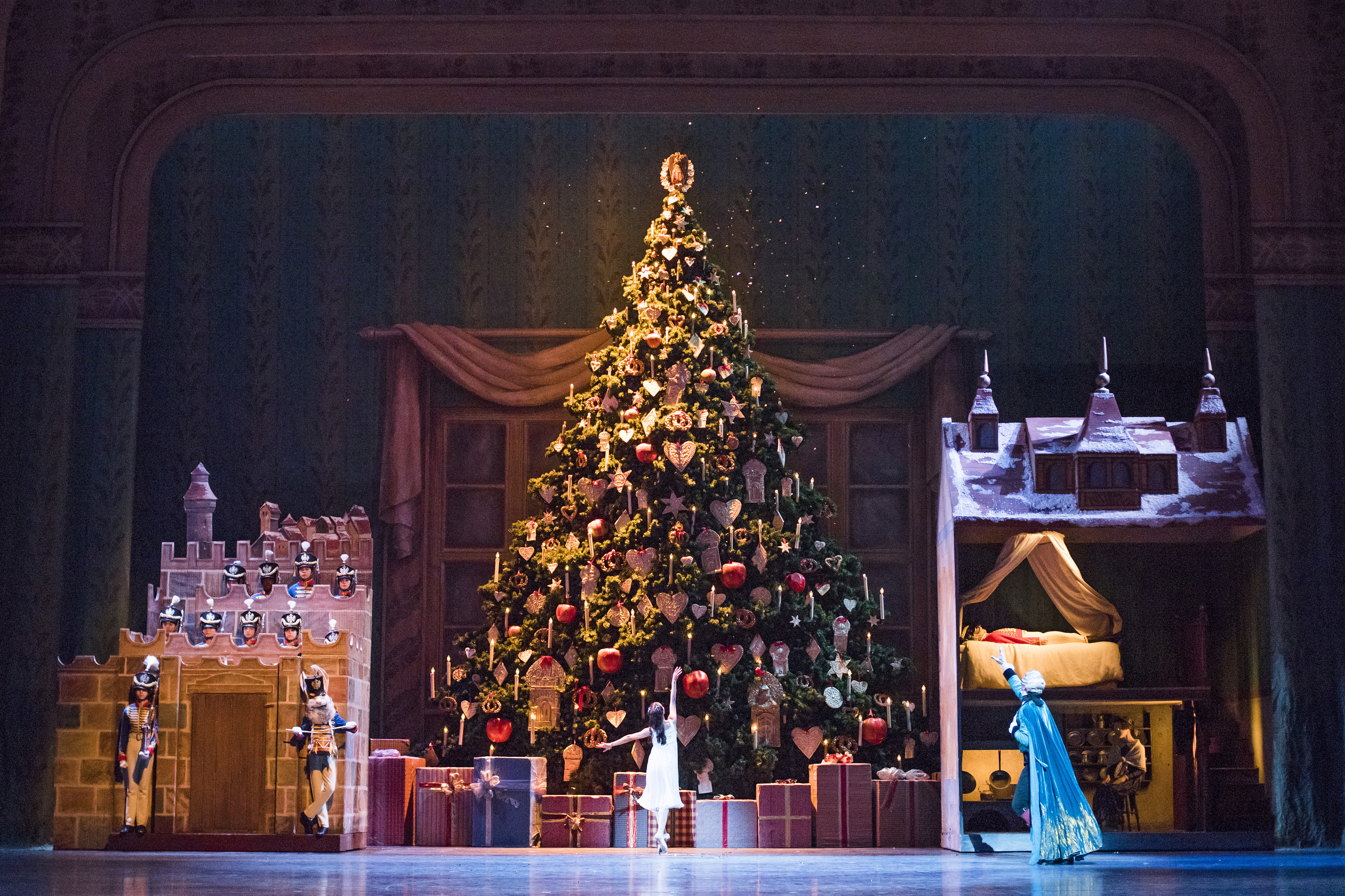 On the Royal Opera House stage, a ballet dancer lifts her arm and faces an enormous Christmas tree that reaches the top of the stage. To the left of the tree a miniature castle is guarded by ballet dancers in military costumes. To the right of the tree, a  nutcracker doll sleeps in a life-size doll house as another dancer in a long blue cape gestures to him.