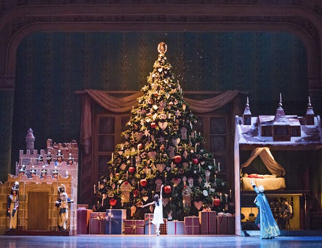 On the Royal Opera House stage, a ballet dancer lifts her arm and faces an enormous Christmas tree that reaches the top of the stage. To the left of the tree a miniature castle is guarded by ballet dancers in military costumes. To the right of the tree, a nutcracker doll sleeps in a life-size doll house as another dancer in a long blue cape gestures to him.