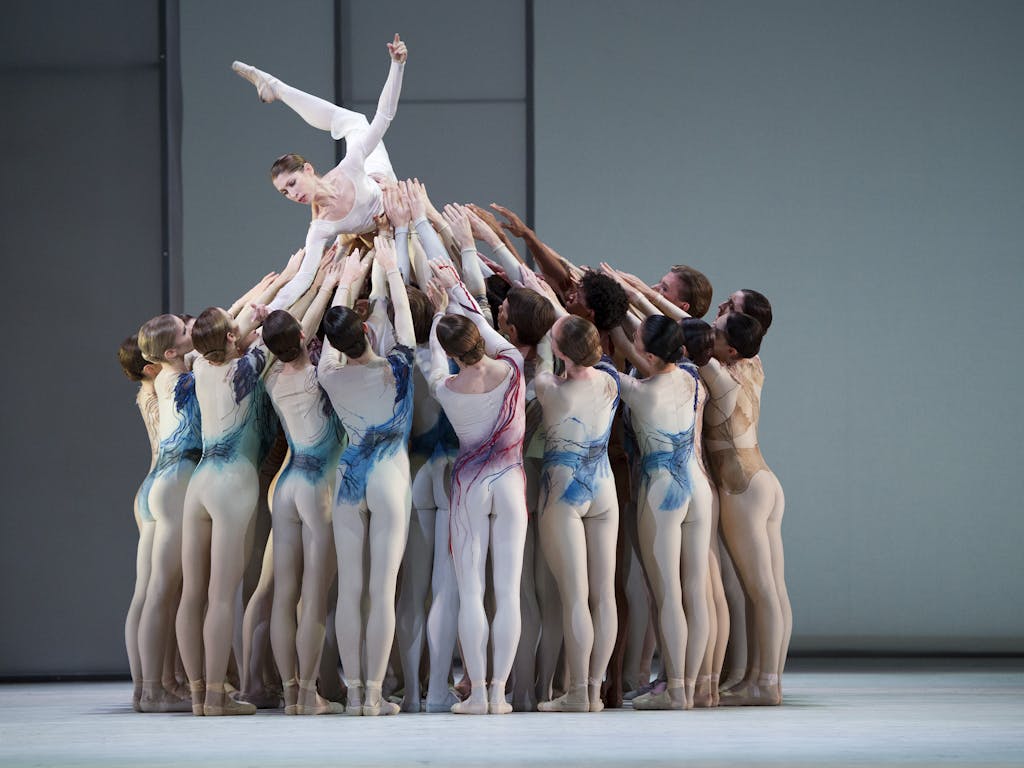 Dancers performing on a light grey stage at the Royal Opera House. A group of dancers in nude bodysuits decorated with splashes of paint in pink, blue and gold are lifting another dancer. The dancer being lifted stretches her hand down toward the group lifting and reaching for her. 