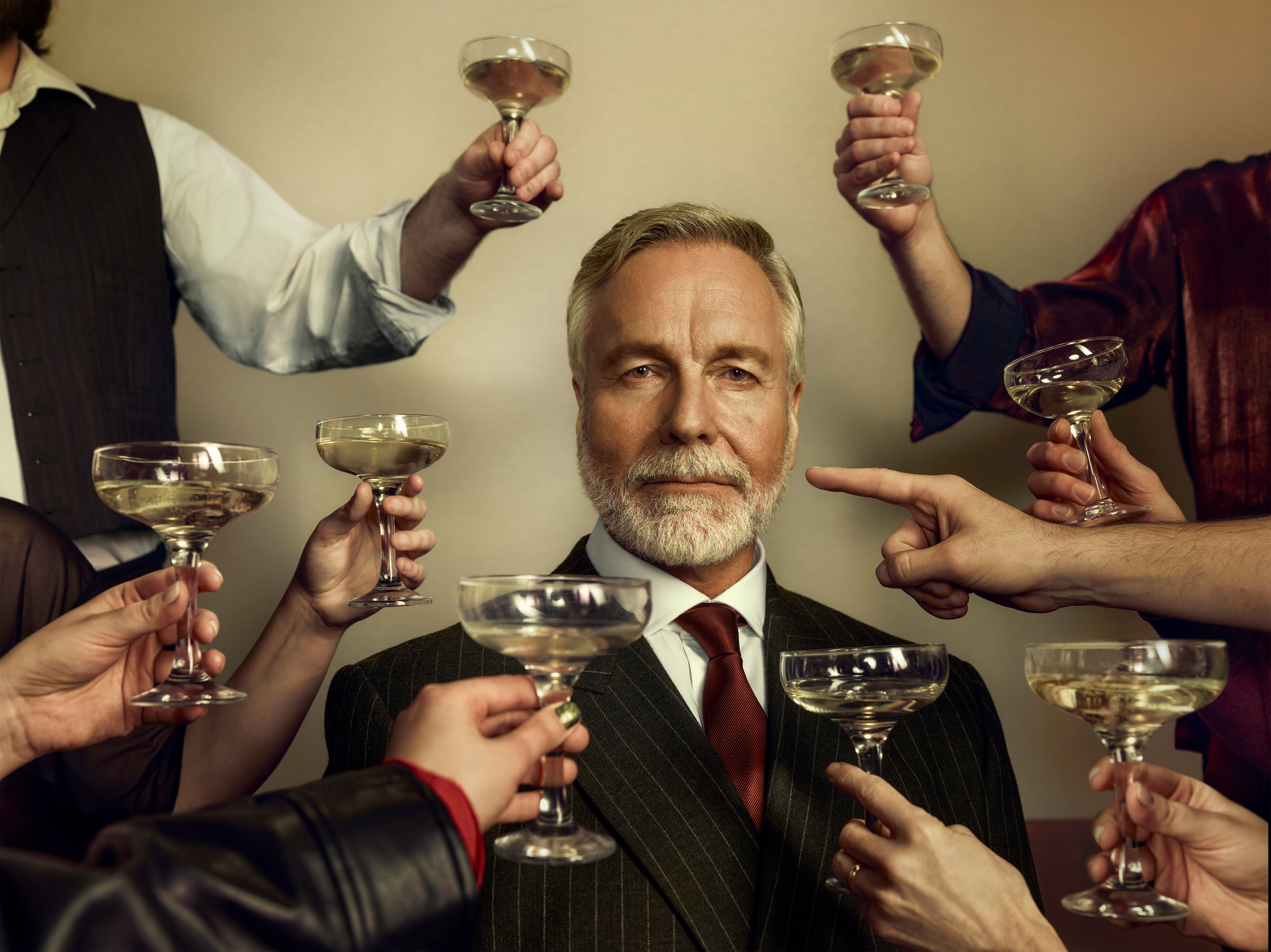 An older man dressed in a sharp suit stares at the viewer as unseen people raise champagne coups in a toast. One arm points a finger at the older man instead.