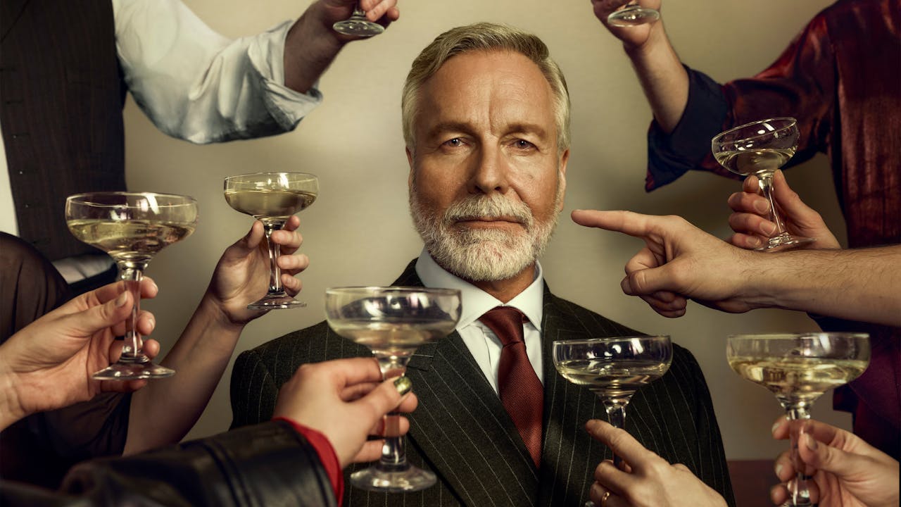 An older man dressed in a sharp suit stares at the viewer as unseen people raise champagne coups in a toast. One arm points a finger at the older man instead.