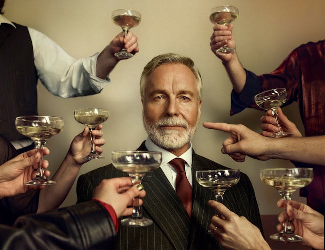 An older man dressed in a sharp suit stares at the viewer as unseen people raise champagne coups in a toast. One arm points a finger at the older man instead.