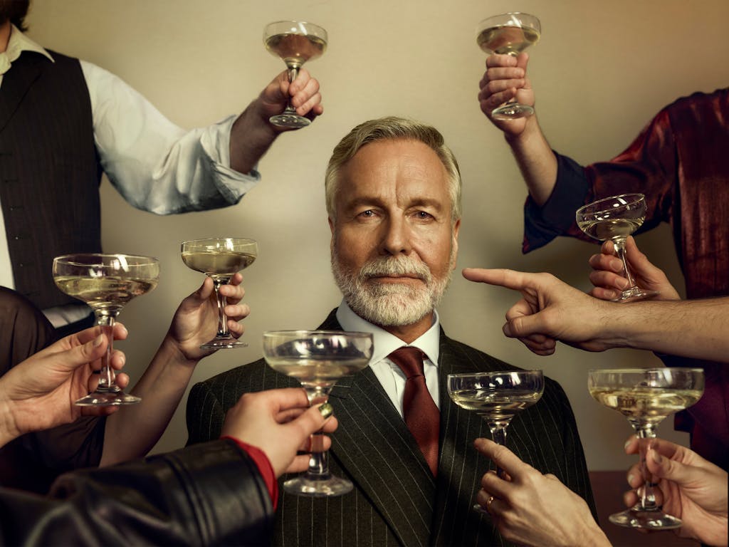 An older man dressed in a sharp suit stares at the viewer as unseen people raise champagne coups in a toast. One arm points a finger at the older man instead.