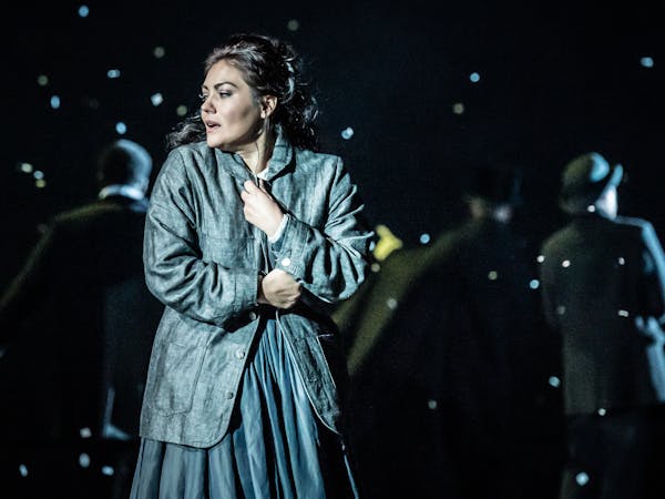 A woman in a dark teal coloured coat holds it close to her body as snow falls around