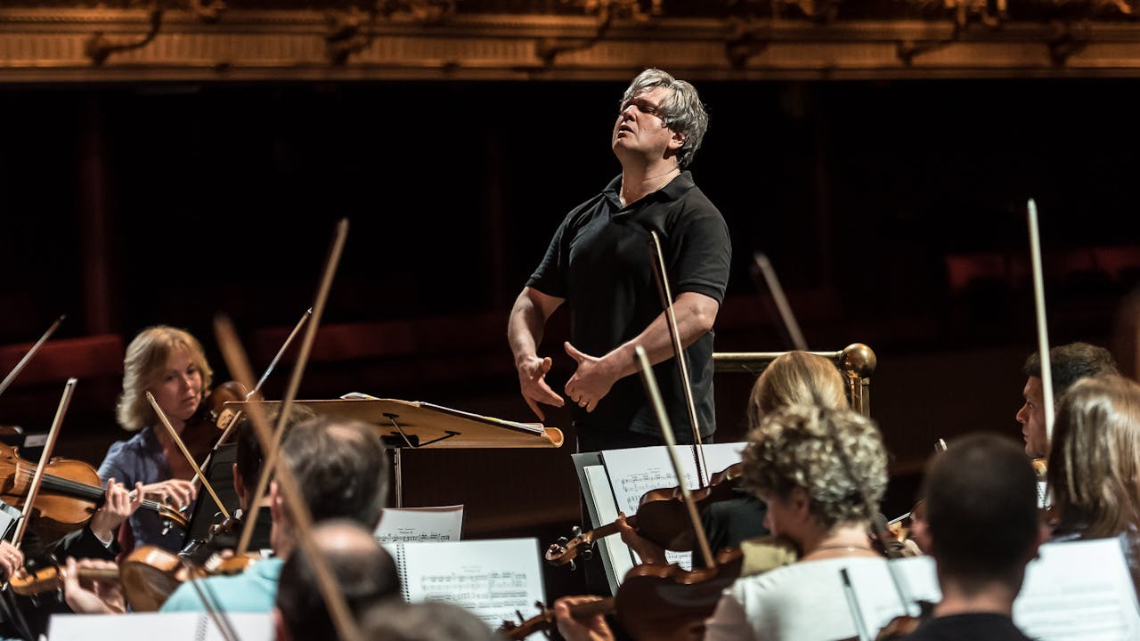 Conductor Antonio Pappano is dressed in black and stands in front of a rapt orchestra. He looks over the top of the musicians and holds his arms in front of him.