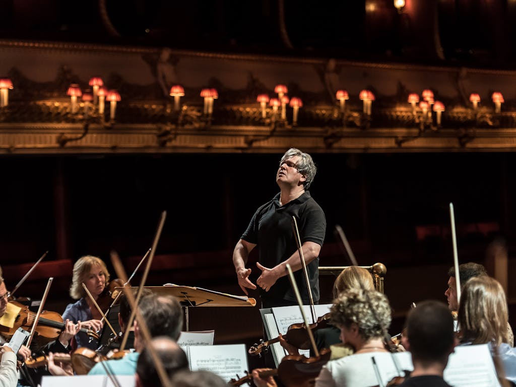 Conductor Antonio Pappano is dressed in black and stands in front of a rapt orchestra. He looks over the top of the musicians and holds his arms in front of him.
