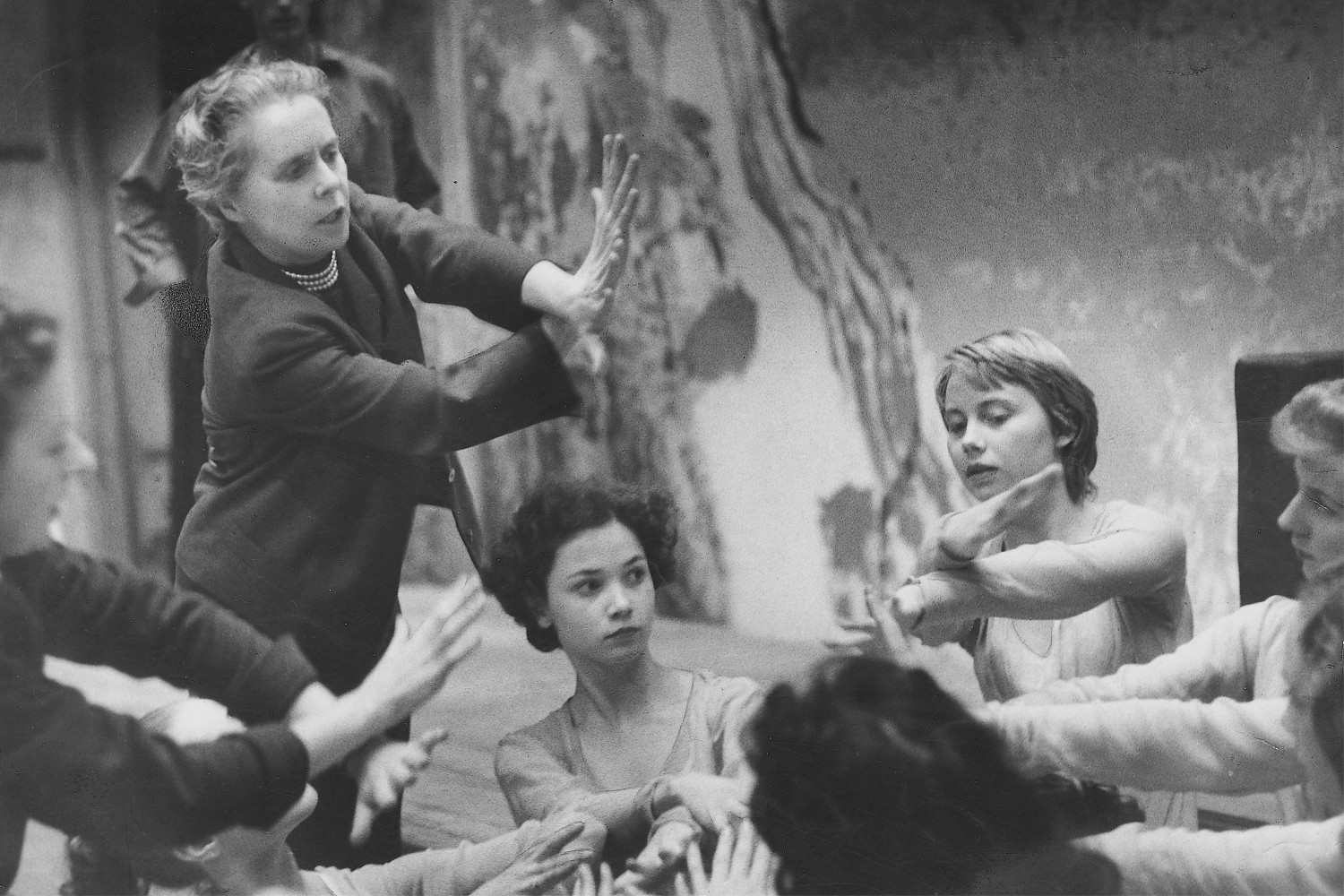 Ninette de Valois standing above a group of dancers as they kneel during a rehearsal. De Valois is demonstrating hand placement to the dancers. 