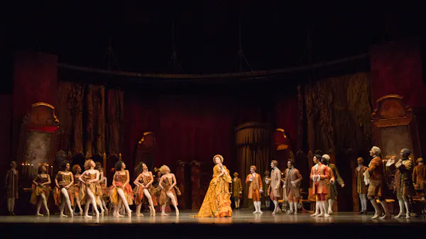 A performance of Manon on the Royal Ballet stage. The set has enormous pillars, and is decorated in a deep red, with heavy curtains and gilded details. A woman clutching a fan smiles at the audience. On one side of the stage, a group of women dressed in French revolution era costumes lift their orange dresses to show their legs. On the other side of the stage, a group of men watch appreciatively.