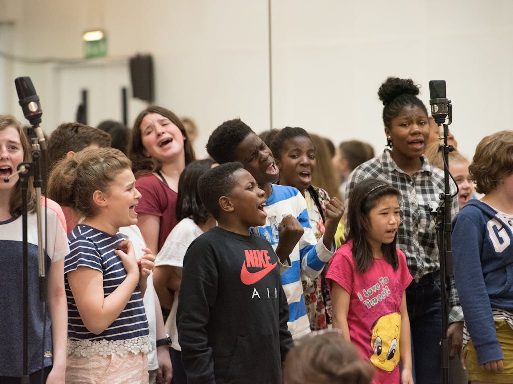 A group of children in casual clothes singing in front of scattered microphones in a crowded room.