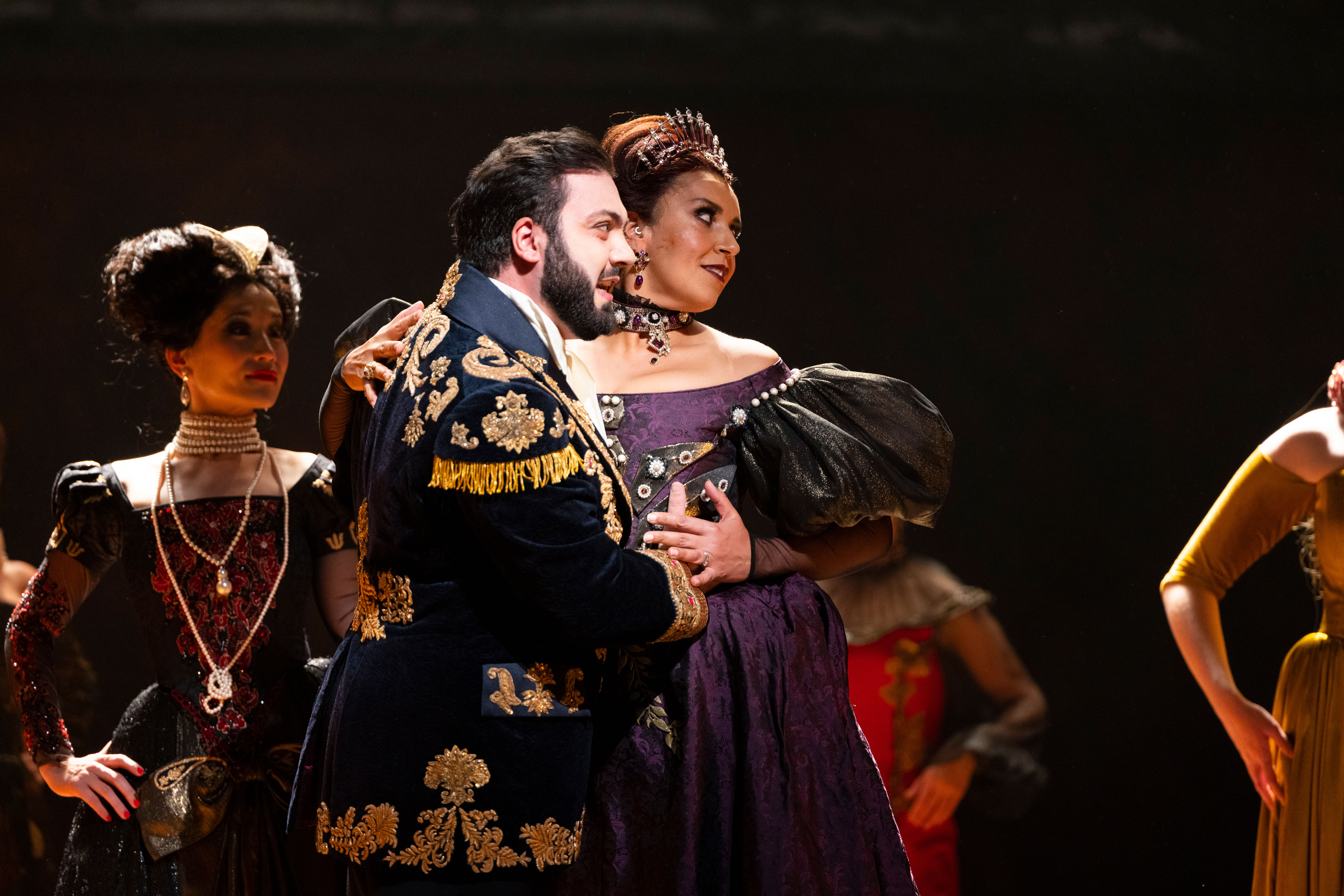 A man in a richly decorate black and gold jacket holds onto a woman in a dark purple dress. 