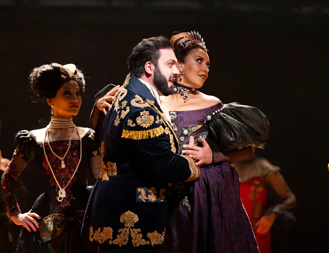 A man in a richly decorate black and gold jacket holds onto a woman in a dark purple dress.