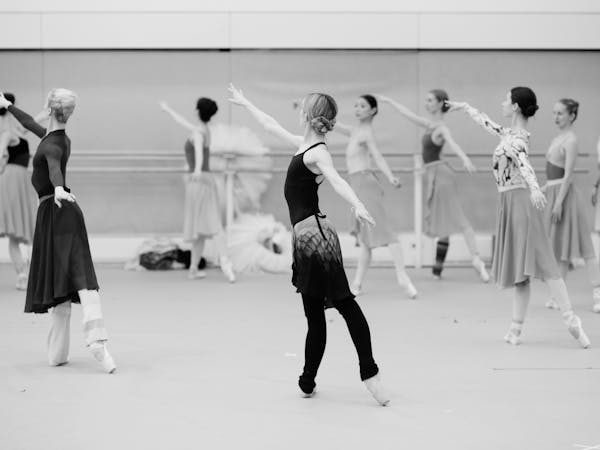 A black and white image of female dancers standing in arabesque, facing the back of the rehearsal studio.
