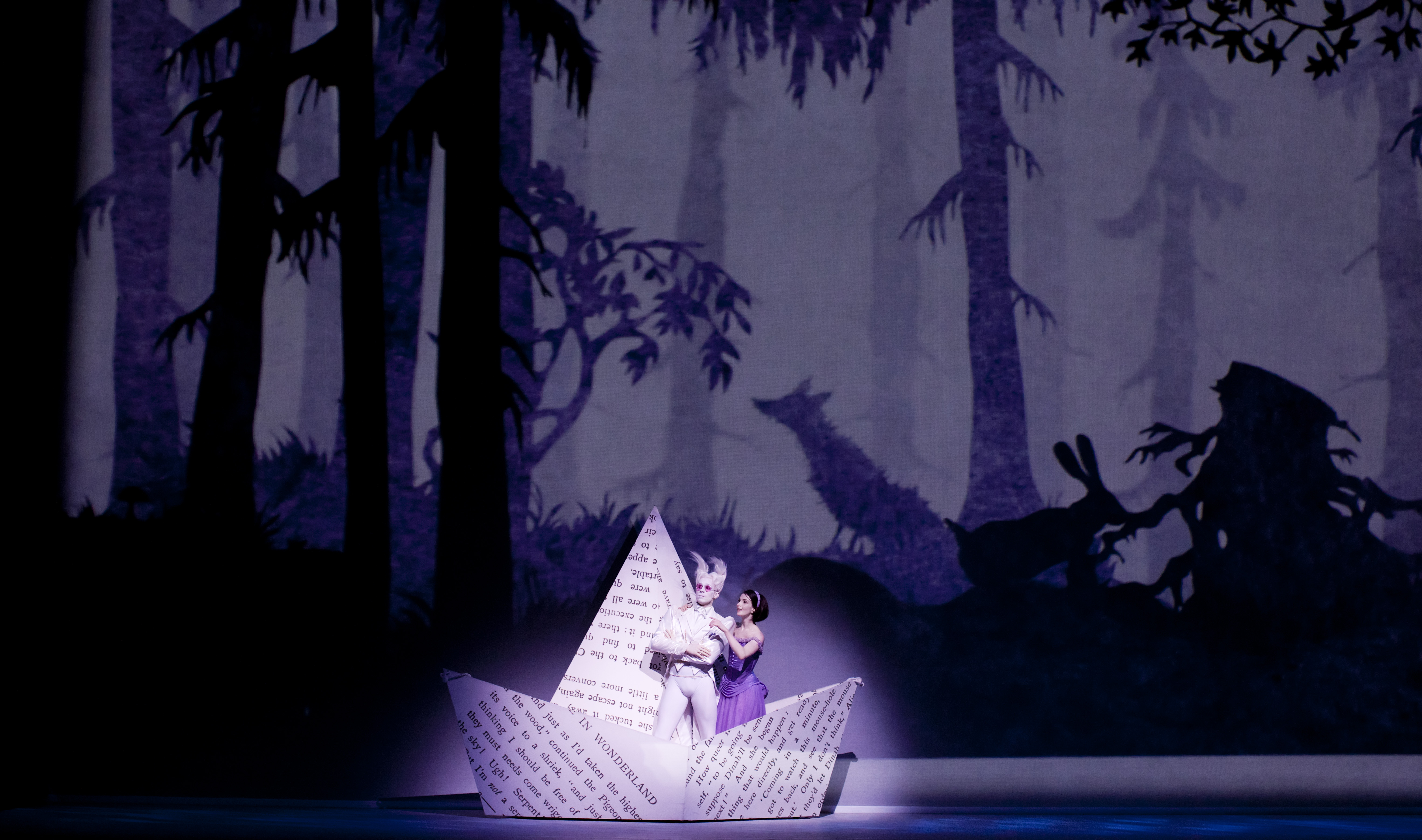 In a production of Alice's Adventures in Wonderland, the stage background is a whimsical woodland scene with animals and trees appearing as a silhouette. A spotlight illuminates Alice, who has brown hair and is wearing a purple dress, and the white rabbit, who has pointed ears and pink sunglasses. They are standing in an enlarged paper boat, folded from a page from the book Alice's Adventures in Wonderland. 