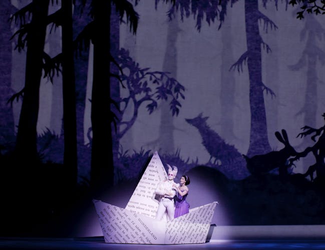 In a production of Alice's Adventures in Wonderland, the stage background is a whimsical woodland scene with animals and trees appearing as a silhouette. A spotlight illuminates Alice, who has brown hair and is wearing a purple dress, and the white rabbit, who has pointed ears and pink sunglasses. They are standing in an enlarged paper boat, folded from a page from the book Alice's Adventures in Wonderland.