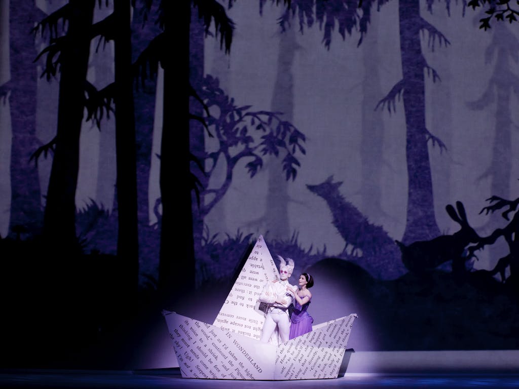 In a production of Alice's Adventures in Wonderland, the stage background is a whimsical woodland scene with animals and trees appearing as a silhouette. A spotlight illuminates Alice, who has brown hair and is wearing a purple dress, and the white rabbit, who has pointed ears and pink sunglasses. They are standing in an enlarged paper boat, folded from a page from the book Alice's Adventures in Wonderland.