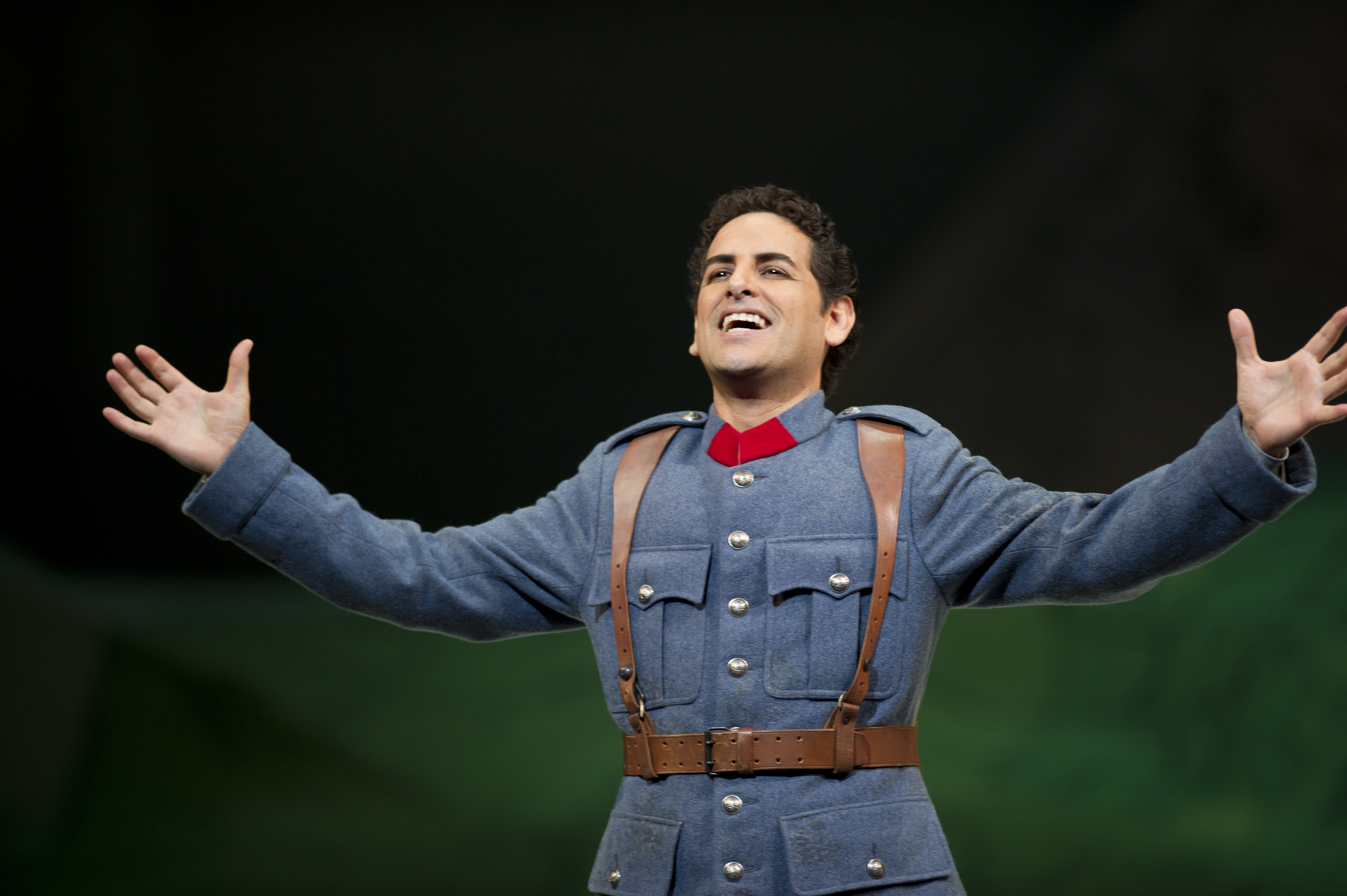 Juan Diego Florez stands arm stretched in the centre of the image. He is smiling broadly. He is wearing a blue military uniform with brown leather belt and braces.