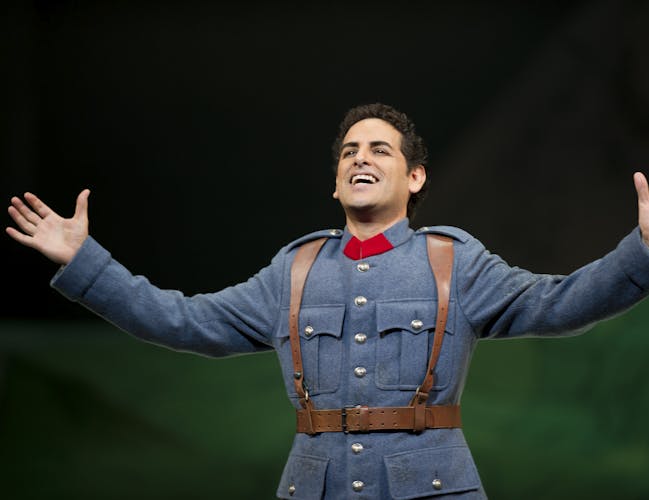 Juan Diego Florez stands arm stretched in the centre of the image. He is smiling broadly. He is wearing a blue military uniform with brown leather belt and braces.