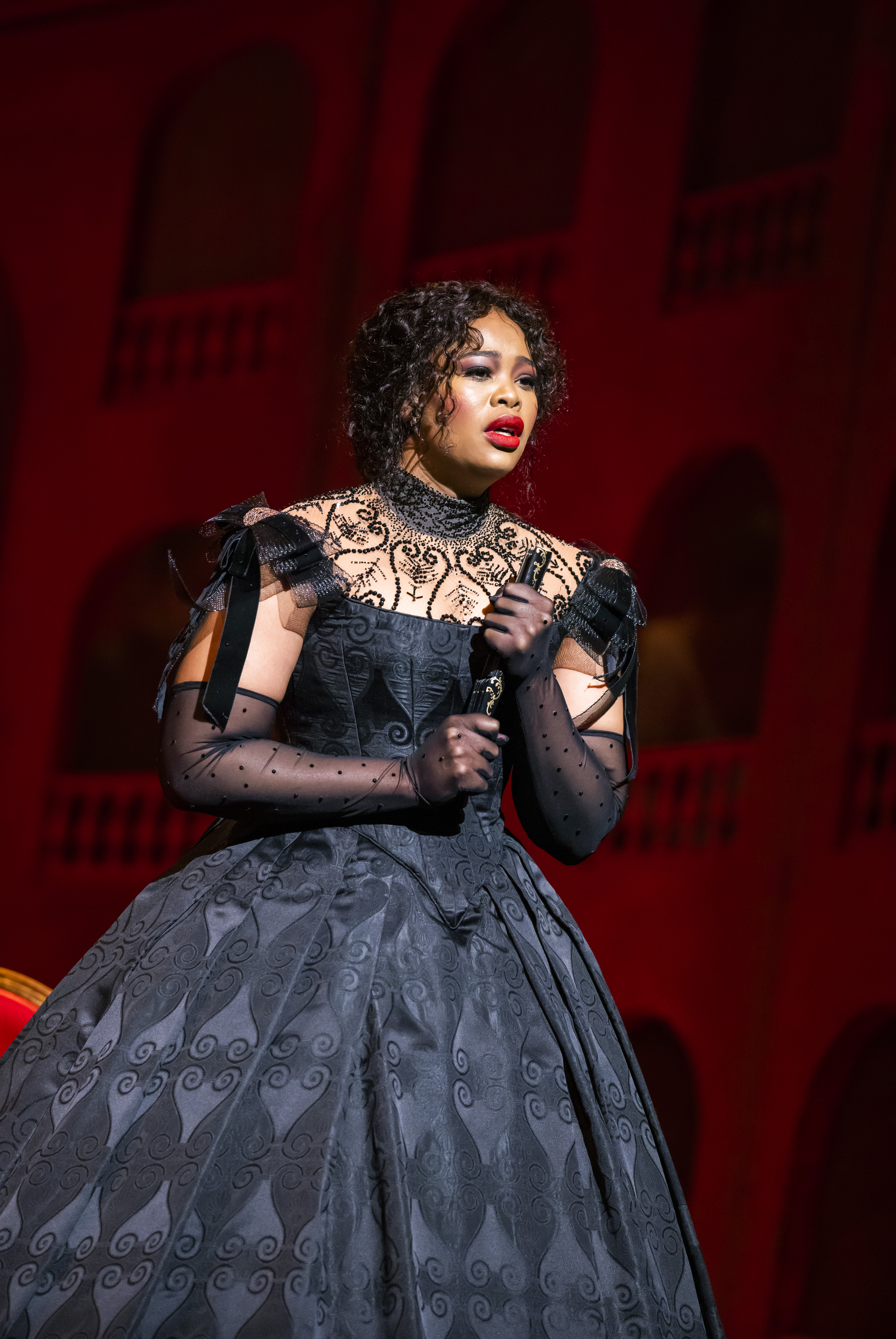 A woman in a black dress holding a fan looks out onto the audience from the stage as she sings.