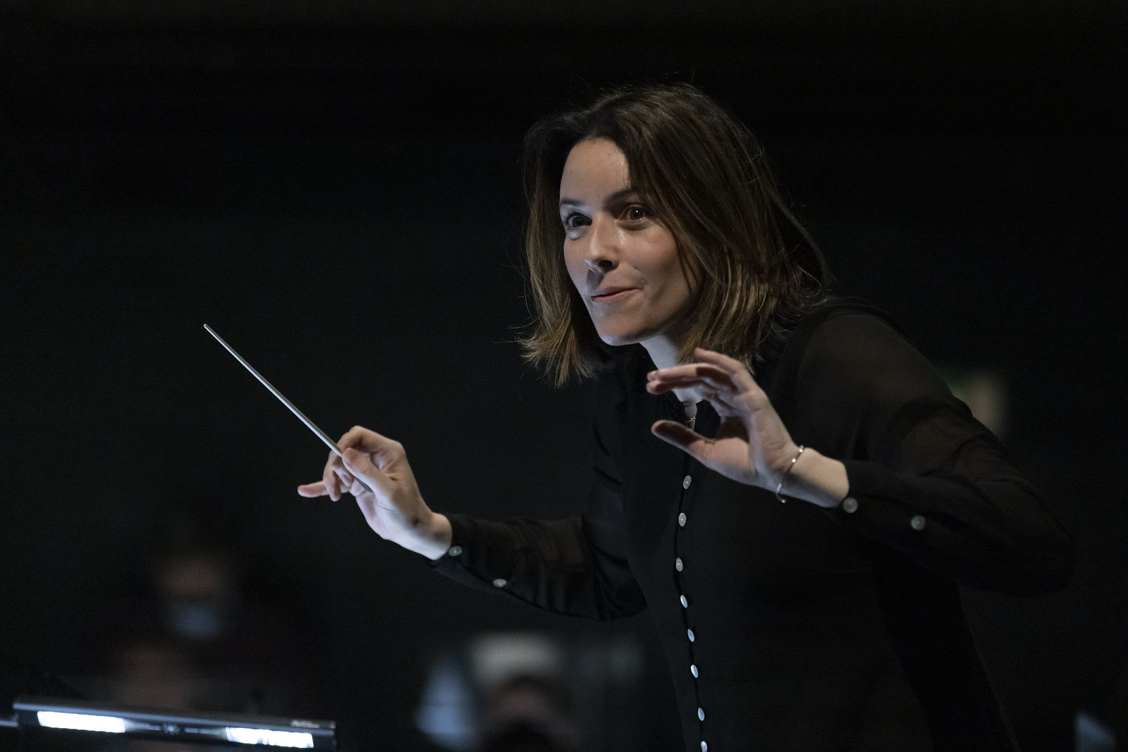 A conductor holds up a baton. Blurred faces in the background represent an orchestra. It is an image of conductor Alondra De La Parra.