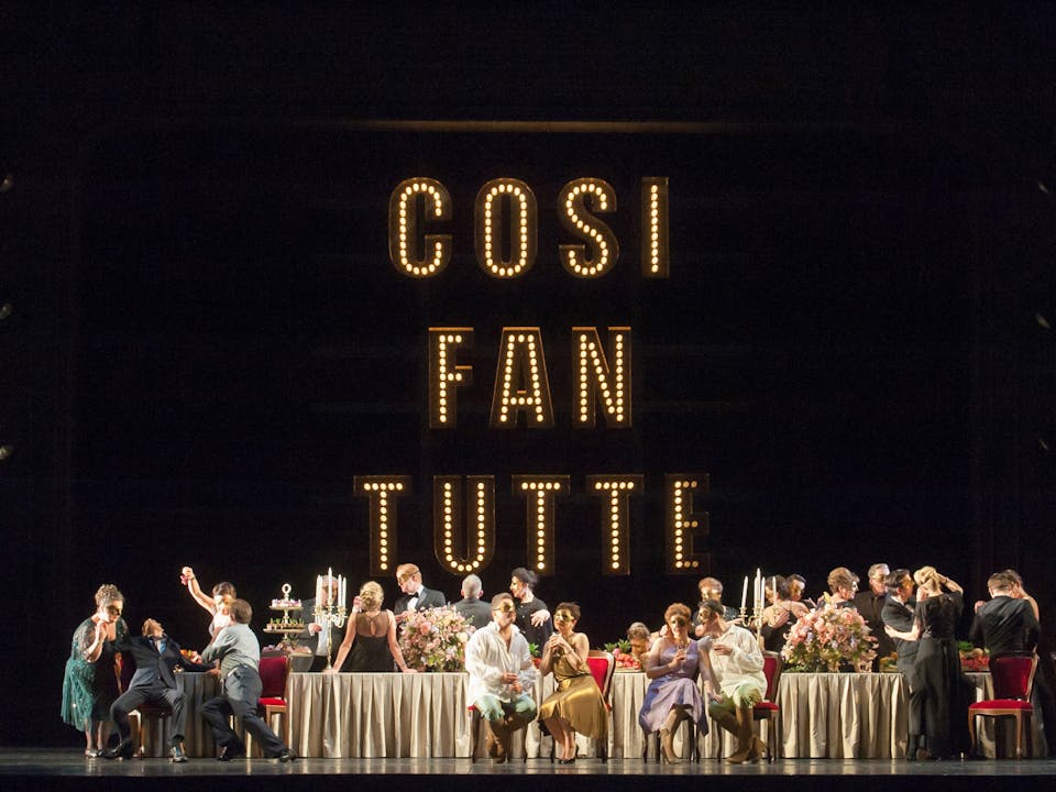 An ensemble cast dressed in their evening best and masquerade masks celebrate around a long table under bright lights that say 'Cosi Fan Tutte' onstage at the Royal Opera House.