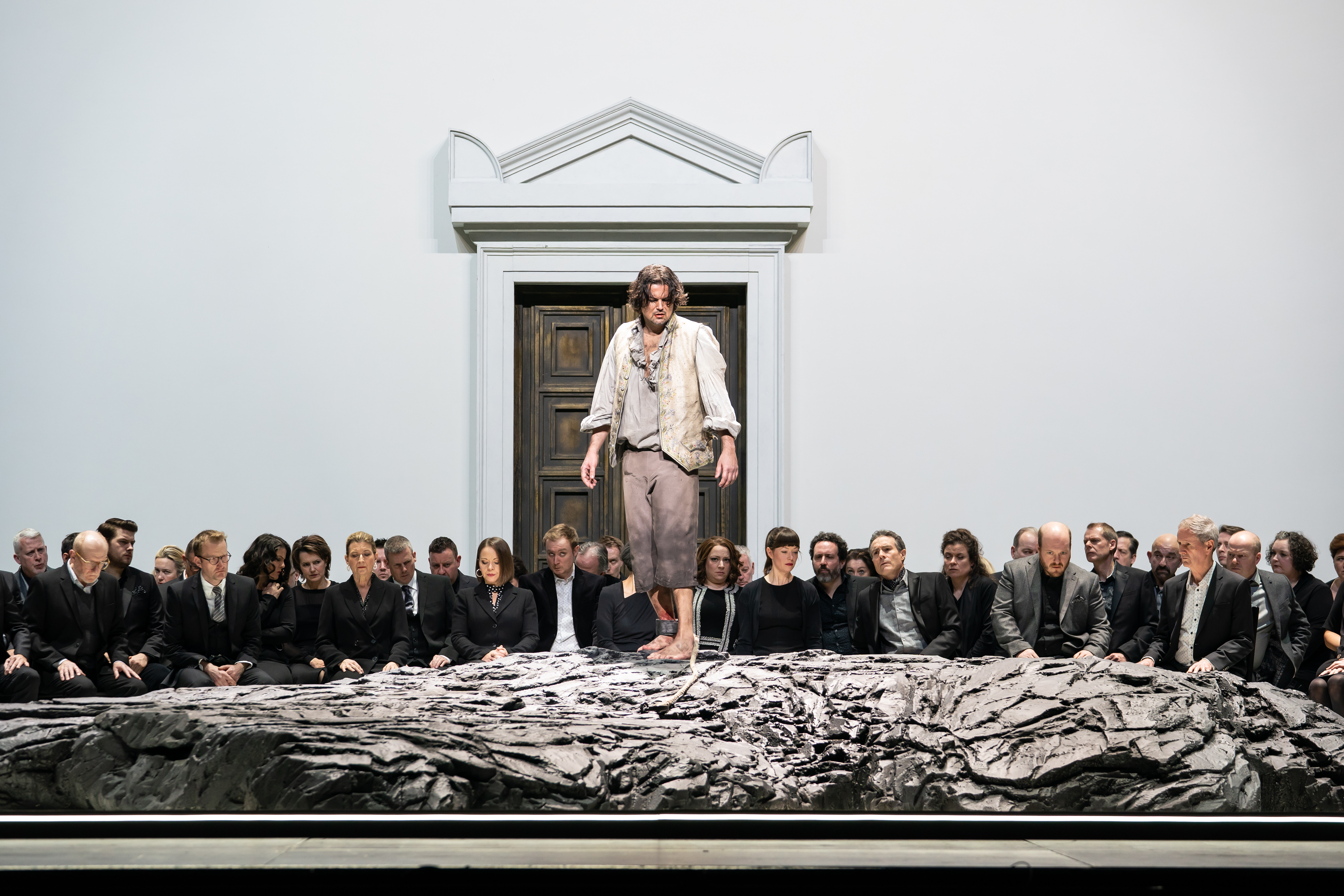 A performer stands on a raised rock with their ankle shackled. Behind them a group of people look on with serious expressions on their face