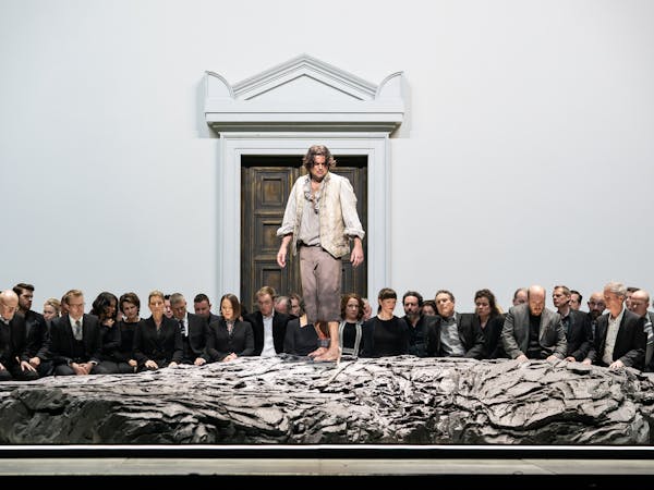 A performer stands on a raised rock with their ankle shackled. Behind them a group of people look on with serious expressions on their face