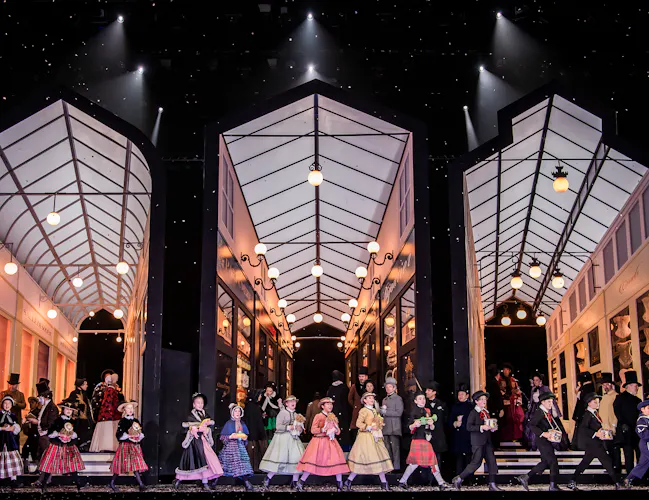 A large set of three arcades appear on stage. In front of the set children hold presents and sing, they are wearing colourful dresses. (2 of 4)
