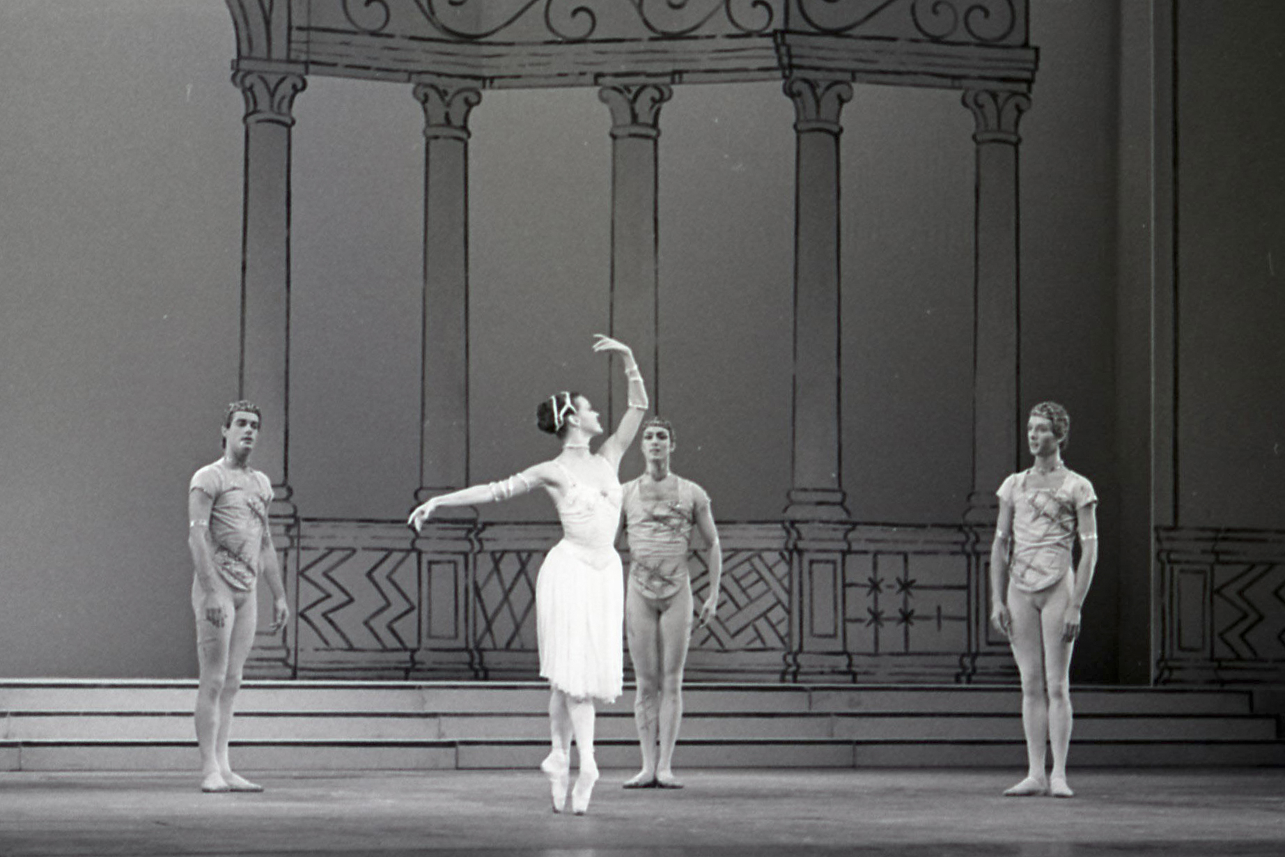 A dancer stands on stage in a white dress. She stands en pointe and raises one arm above her head. Behind her are columns. Three male dancers stand in front of the columns arms at their sides (2 of 8)
