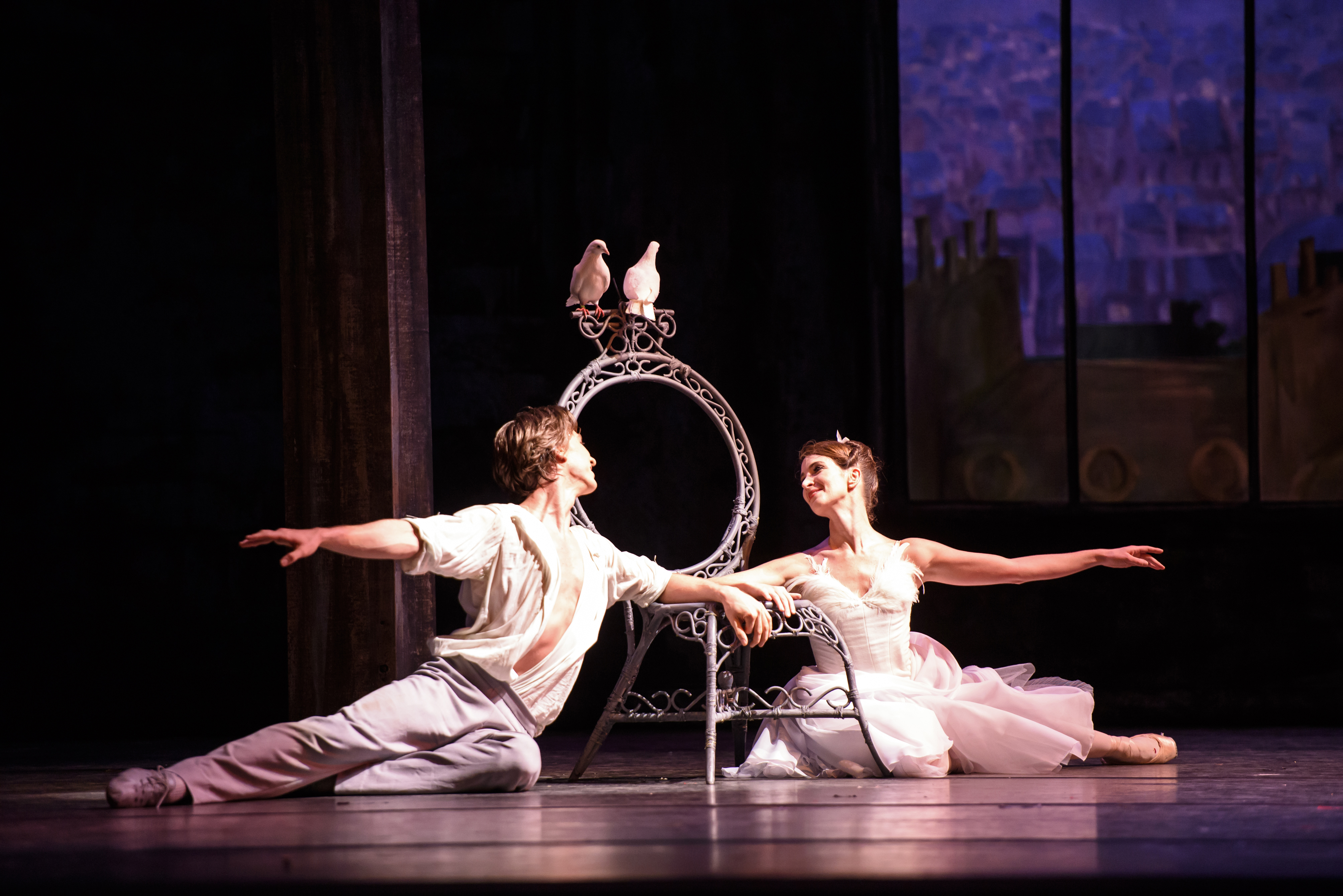 Two dancers sit on the floor, reaching to each other. In the background two pigeons sit on a chair.