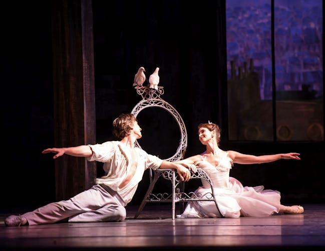 Two dancers sit on the floor, reaching to each other. In the background two pigeons sit on a chair.