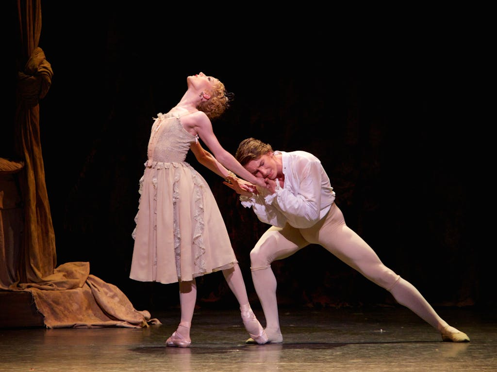 Ballet dancer Sarah Lamb is wearing a pale pink dress with frills and ribbons. Her blonde hair is pinned up with a sparkling hair piece. She is facing upwards, away from dancer Vadim Muntagirov who is clasping her hands. 