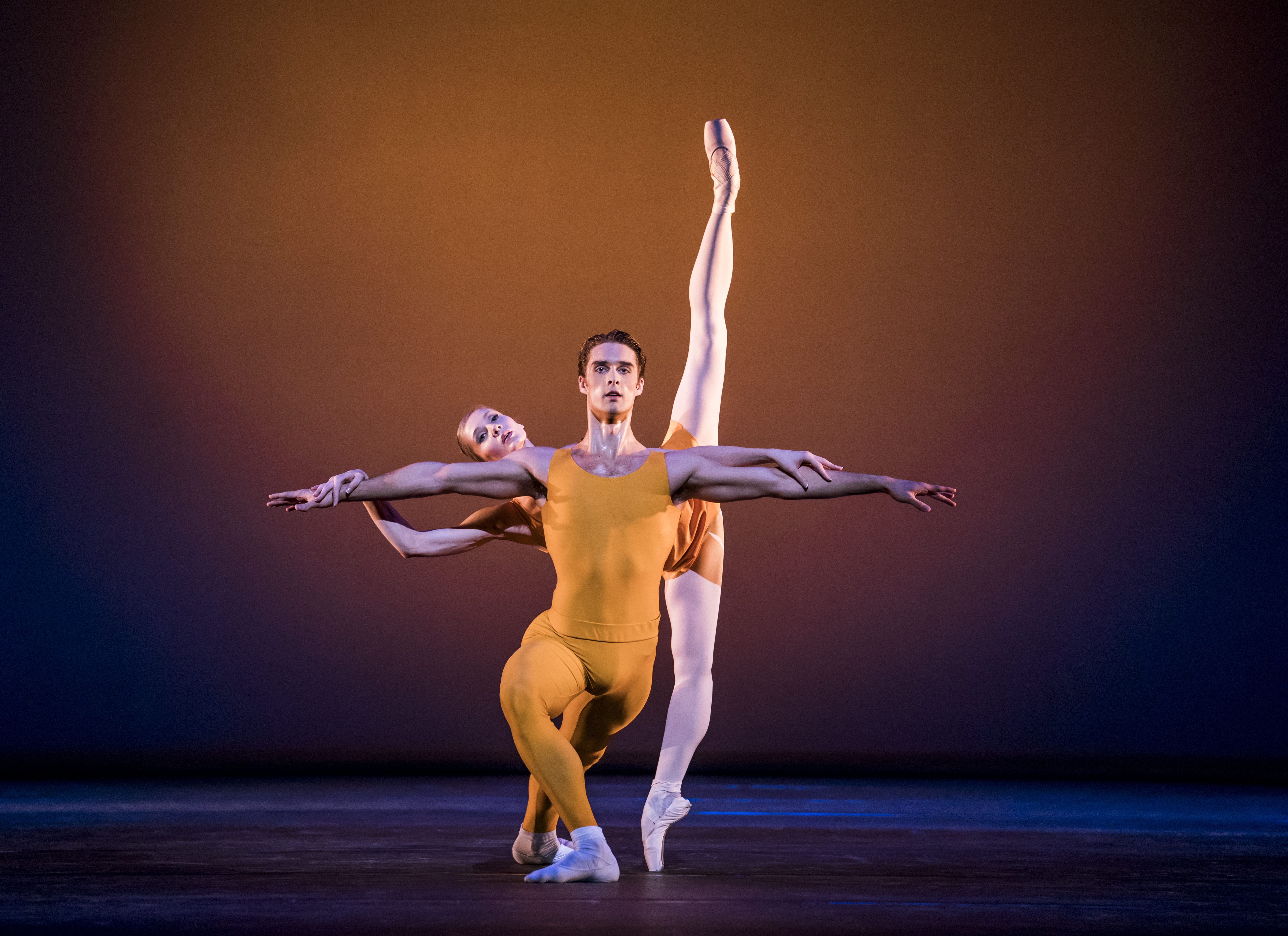Two dancers wearing yellow leotards appear on stage. The dancer in front crouches with one knee bent in front of him and arms outstretched to the side. Behind him the other dancer has her legs in the splits in a vertical line from the floor, she balances her body on the man's back. (1 of 3)